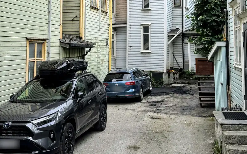 Grijze SUV met een zwarte dakkoffer in een straat met houten huizen en bergen op de achtergrond