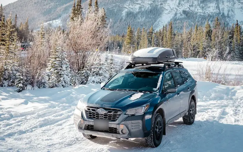 Blauwe SUV met een besneeuwde dakkoffer op een sneeuwweg voor een berg, kenteken onherkenbaar gemaakt